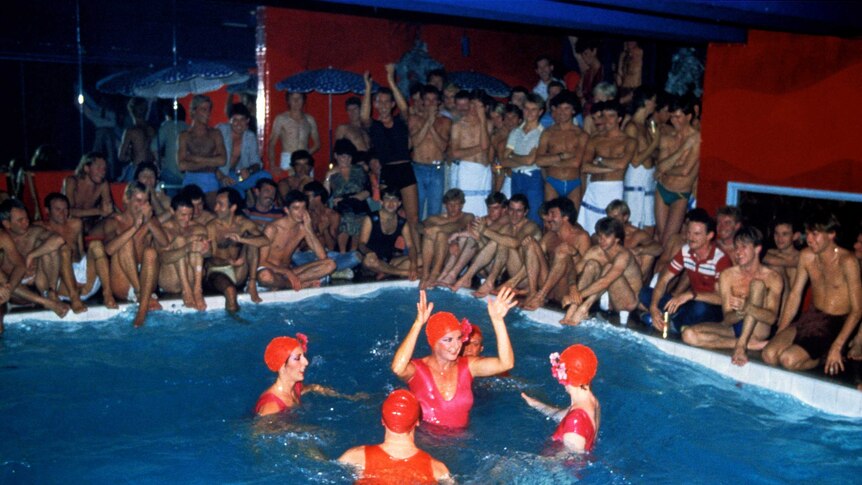 A party in the 1970s with a pool filled with synchronised swimmers at its centre, crowds of shirtless men surround