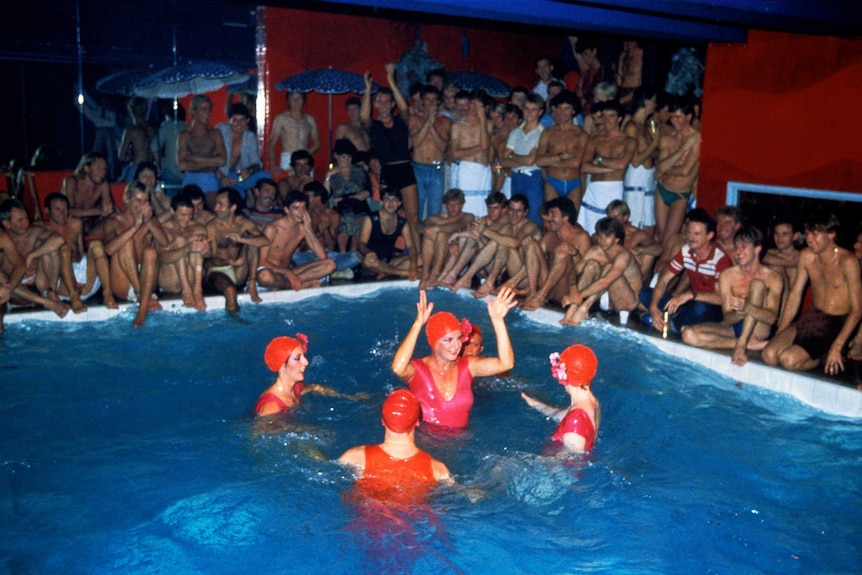 A party in the 1970s with a pool filled with synchronised swimmers at its centre, crowds of shirtless men surround
