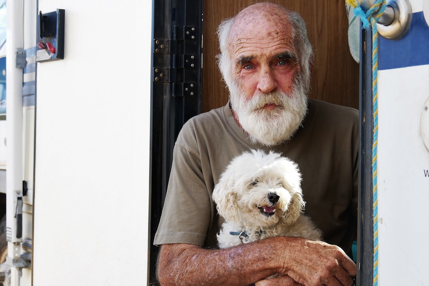 a man sitting in the door of a caravan, holding a small white dog