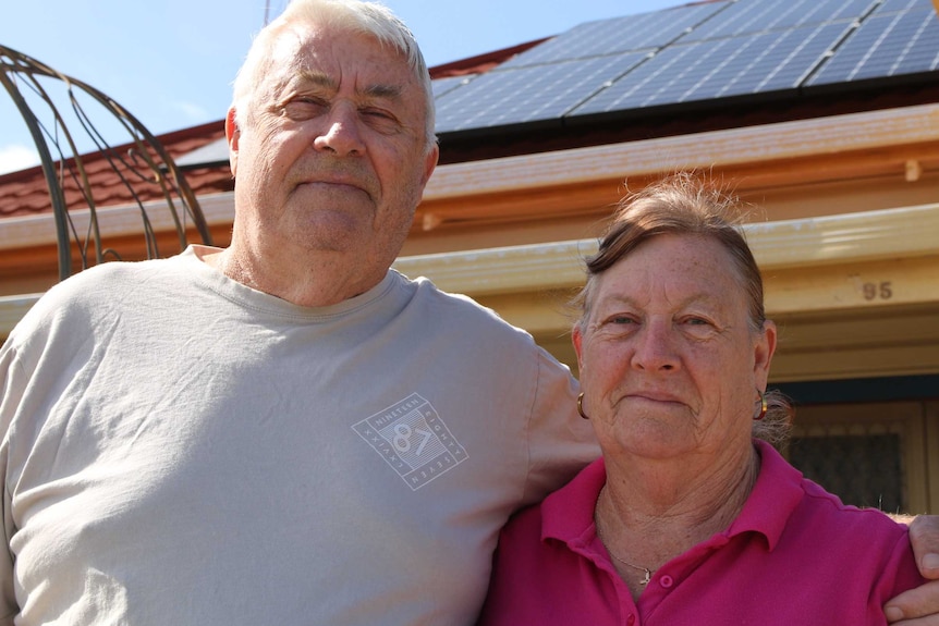 An older couple pose together outside their home which has solar panels mounted on the roof.