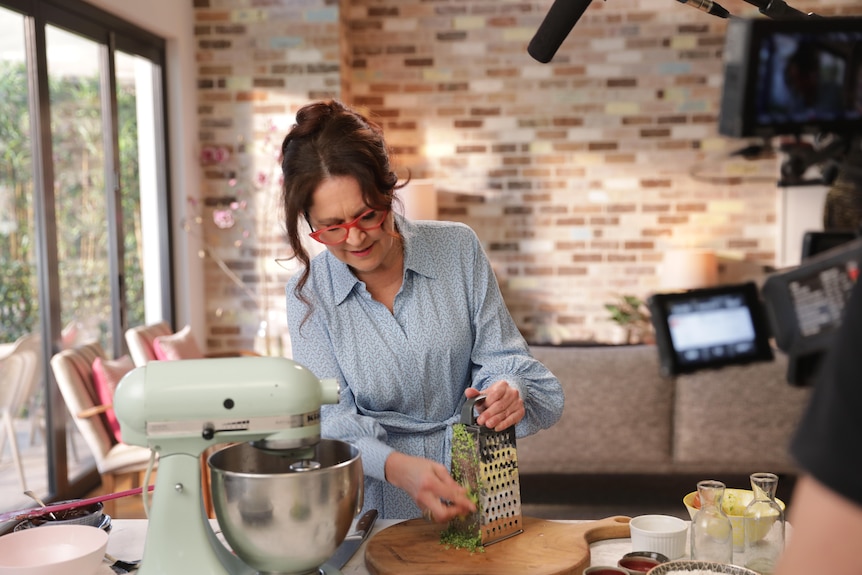 Woman in a kitchen using a grater with a camera filming.