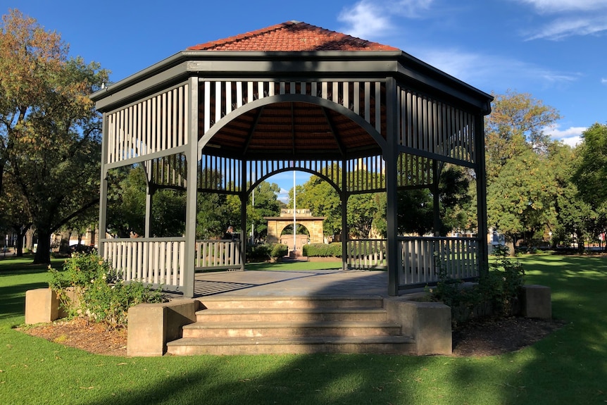 A wooden rotunda in a park with a memorial arch behind