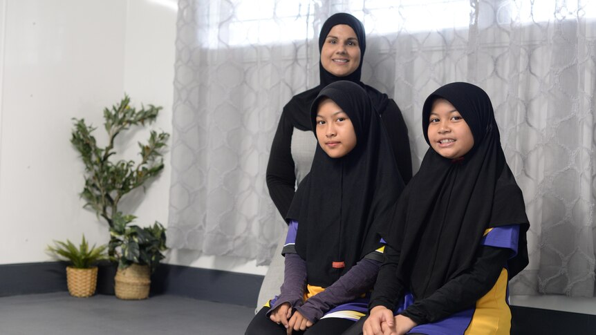 A woman and two primary school-aged girls wearing hijabs kneel on the floor of a well-lit room.