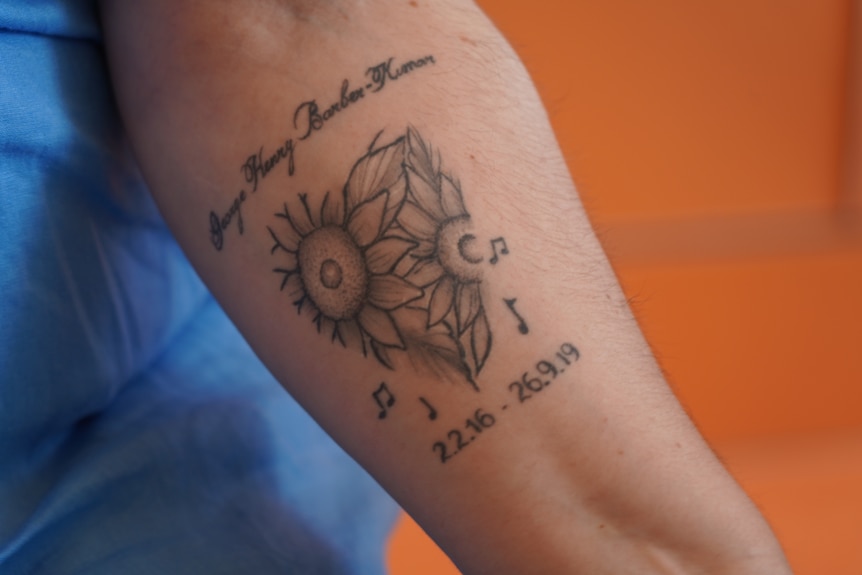 A tattoo of two flowers. Above it cursive writing says George Henry Barber-Kumar. Underneath is musical notes.