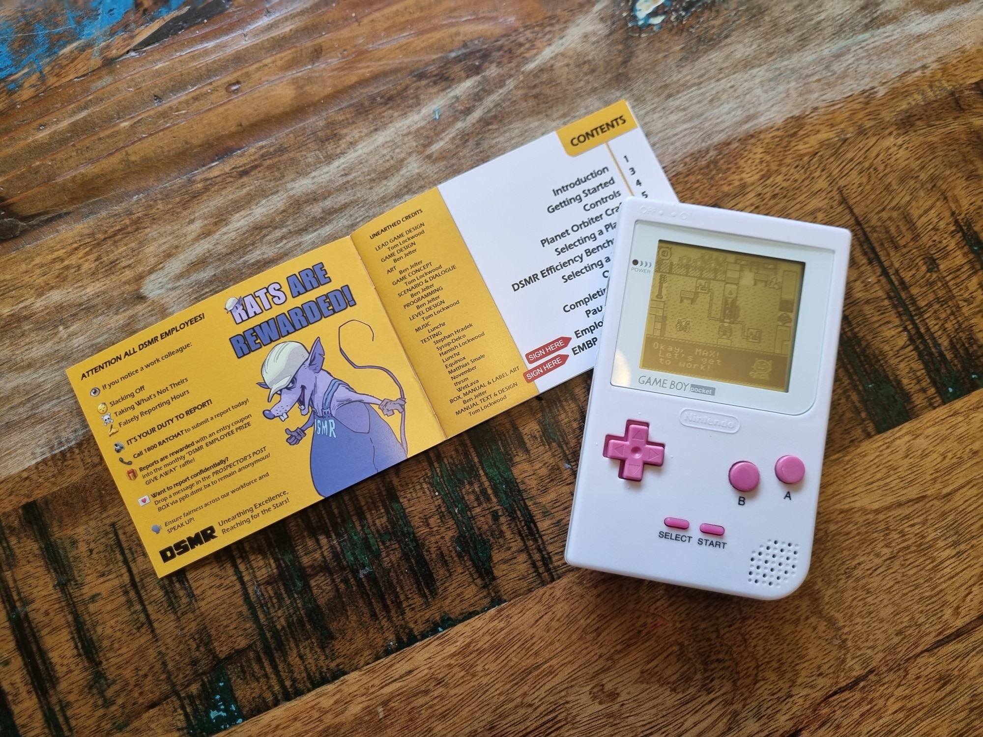 A game boy console sits on a table displaying a game on it's screen with an open game manual behind it. 