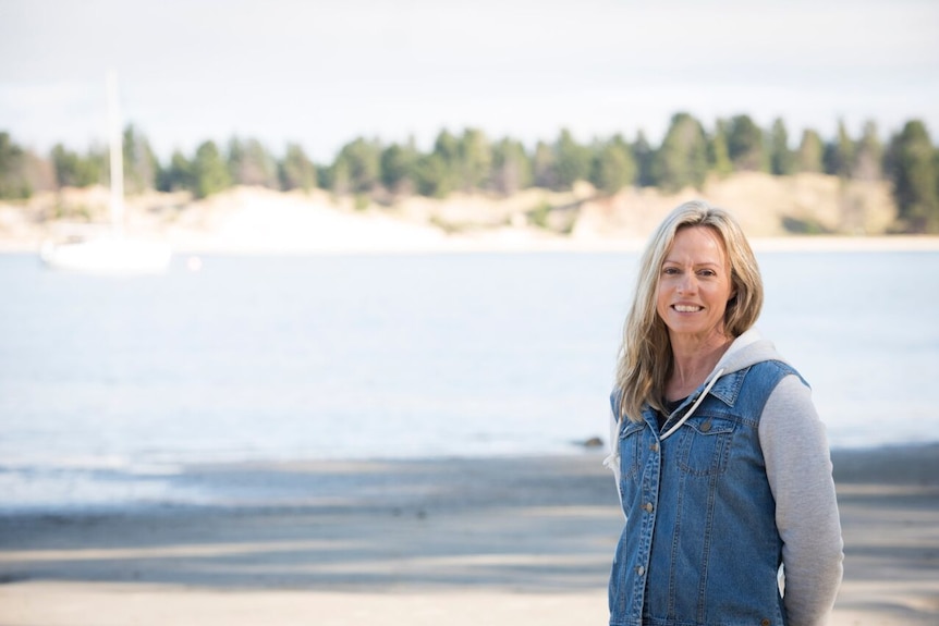 A woman with blonde hair, wearing a denim top smiling on a beach. Christine Gaby's career has advanced in regional Tasmania.