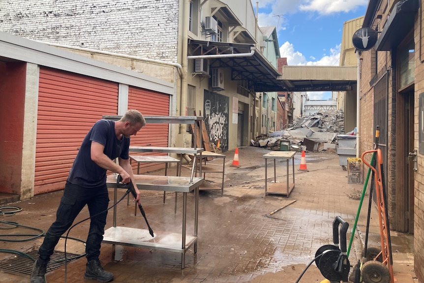 A man stands outside pressure cleaning a stainless steel kitchen bench. Piles of rubbish are in the background.