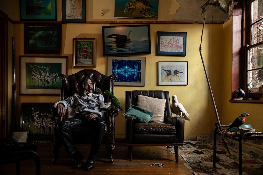 A man in white inside a room, sitting on arm chair surrounded by framed photographs and parrots.