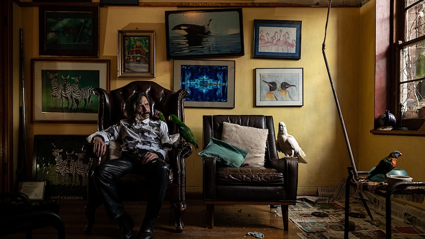 A man in white inside a room, sitting on arm chair surrounded by framed photographs and parrots.