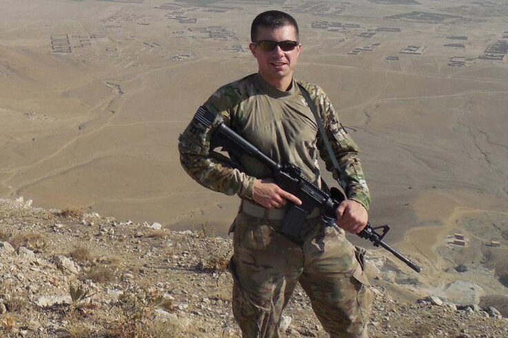 Pete Buttigieg wearing army fatigues and clutching a rifle in an arid location