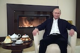 Vladimir Putin sitting by a tea pot and cakes with a fireplace behind him