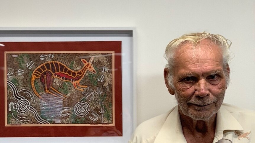 A man in his seventies wears a cream shirt and stands inside next to a re-based indigenous dot painting.