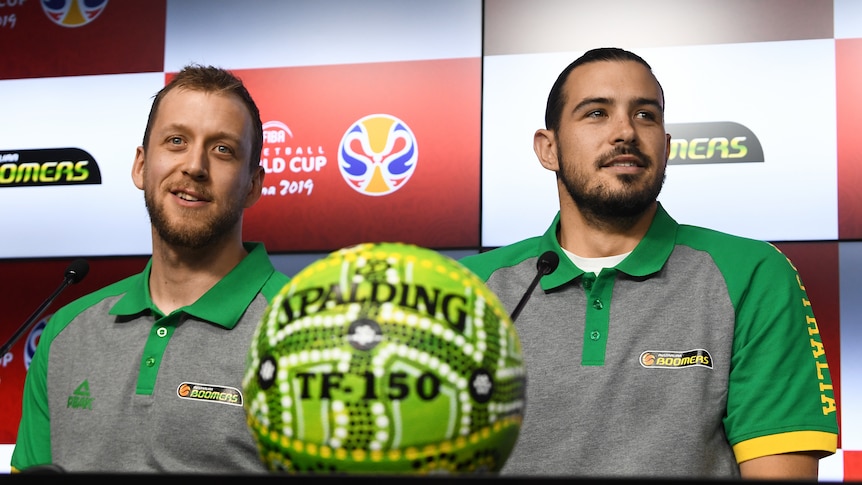 Joe Ingles and Chris Goulding sit next to each other in front of an advertising backdrop with a basketball in front of them