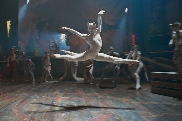 A cream CGI cat with woman's face performs a grand jete ballet movement surrounded by cats in a theatre hall with wooden floor.