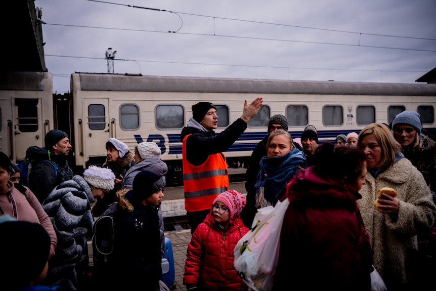 A crowd of people on a train station platform in winter clothes and holding luggage are marshalled by a man in a high-viz vest.