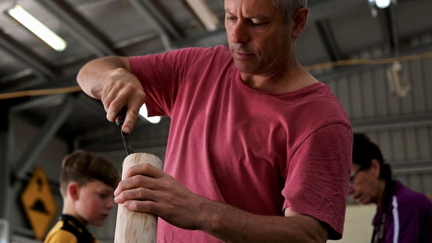 A grey-haired man uses a chisel as he works on a didgeridoo.