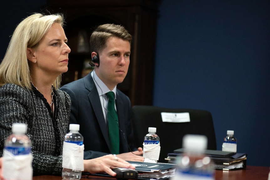 Miles Taylor sits next to a woman at a table. He has an earpiece on.