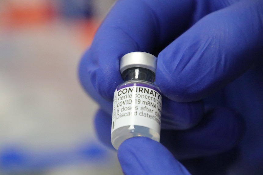 A gloved hand holds a small vial filled with a COVID vaccine.