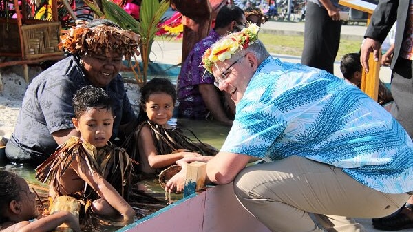 Scott Morrison crouches and smiles next to children who are floating in water.