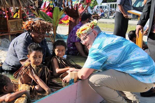 Scott Morrison crouches and smiles next to children who are floating in water.