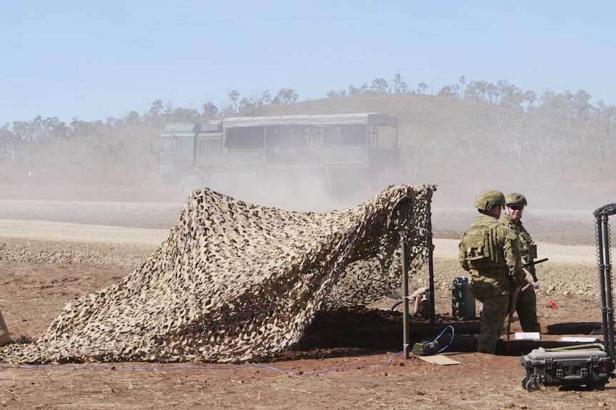 Two men in army uniforms sit on chairs guarding the entry to a military training area on the side of a dirt road