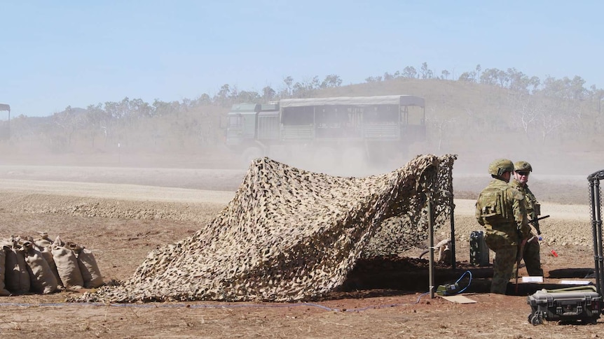 Two men in army uniforms sit on chairs guarding the entry to a military training area on the side of a dirt road