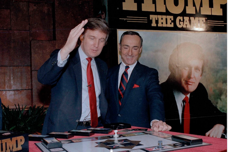 A young Donald Trump gestures standing next to another man in a suit, looking down at a board game