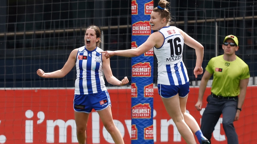 A Kangaroos AFLW player roars in celebration with arms spread, as her teammate points after a goal.