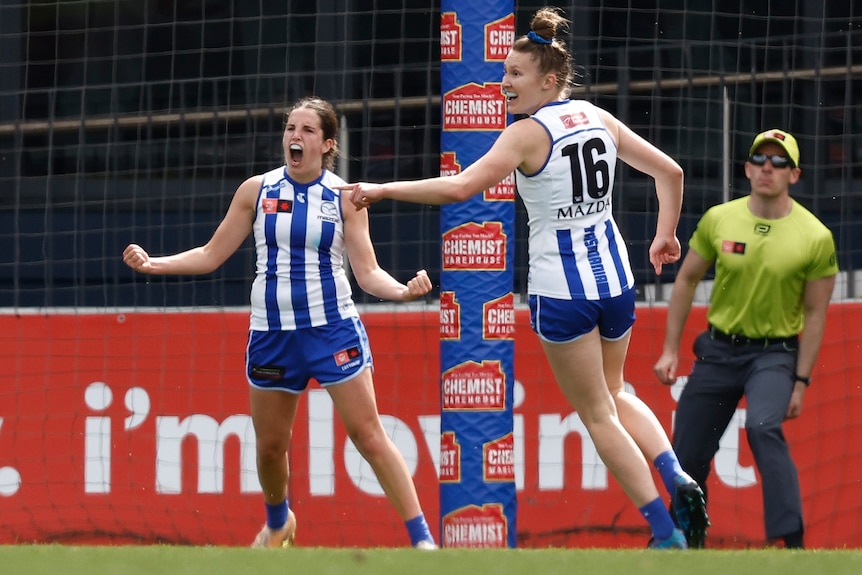A Kangaroos AFLW player roars in celebration with arms spread, as her teammate points after a goal.