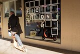 A man rides a scooter past a window that has a sign reading "Fiction".