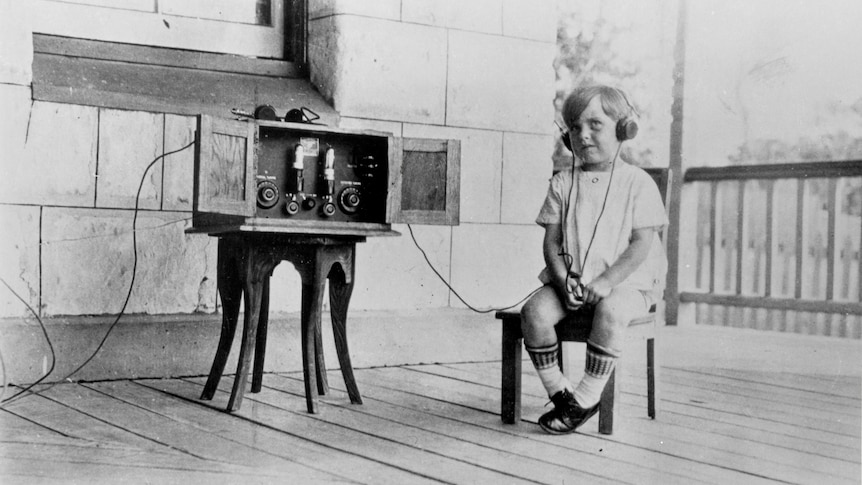 Black and white grainy photo of small child sitting on wooden stool with headphones on, sitting next to old radio in wooden box.
