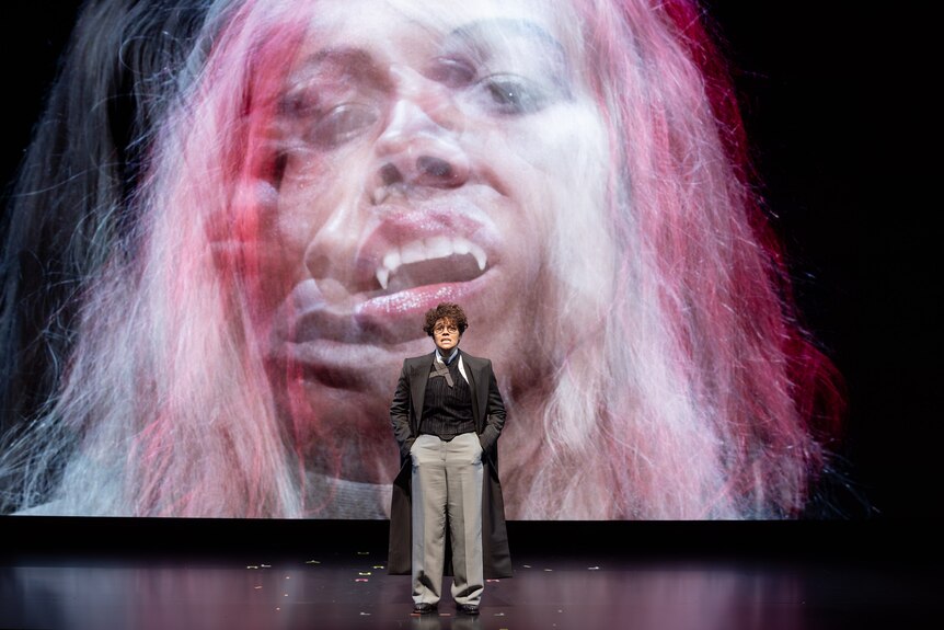 On stage, Zahra Newman, in a wig and glasses, stands, hands in pockets. On a screen behind her is a video of her as Dracula.