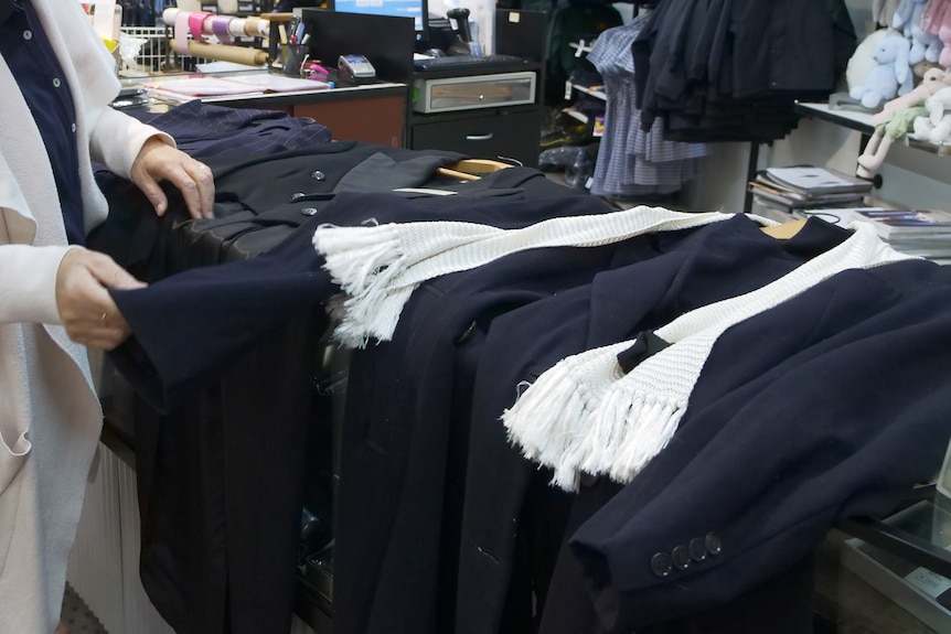 two navy blue and one black coat lying flat on a shop counter. A woman in a white coating is sorting through them. 