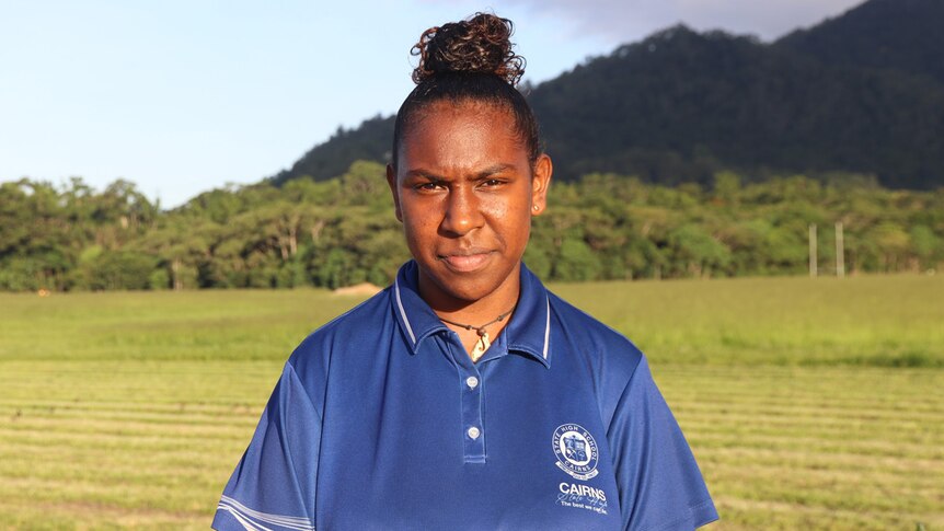 An Indigenous teenage girl in a sports polo shirt stands in front of a field with a mountain in the background.
