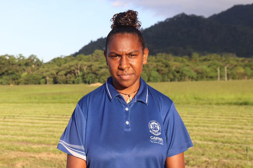 An Indigenous teenage girl in a sports polo shirt stands in front of a field with a mountain in the background.