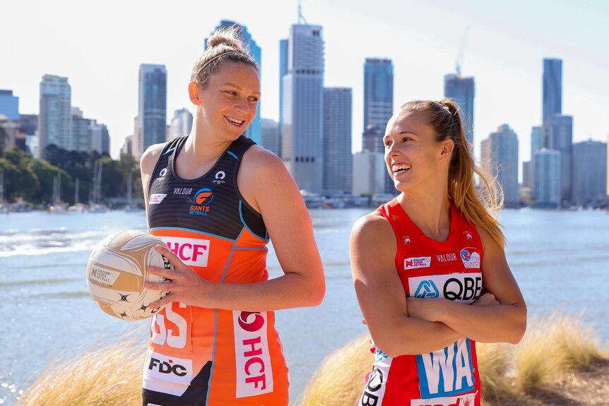Jo Harten stands tall, holding the Grand Final ball, smiling back-to-back at Paige Hadley, in front of a Brisbane sky line