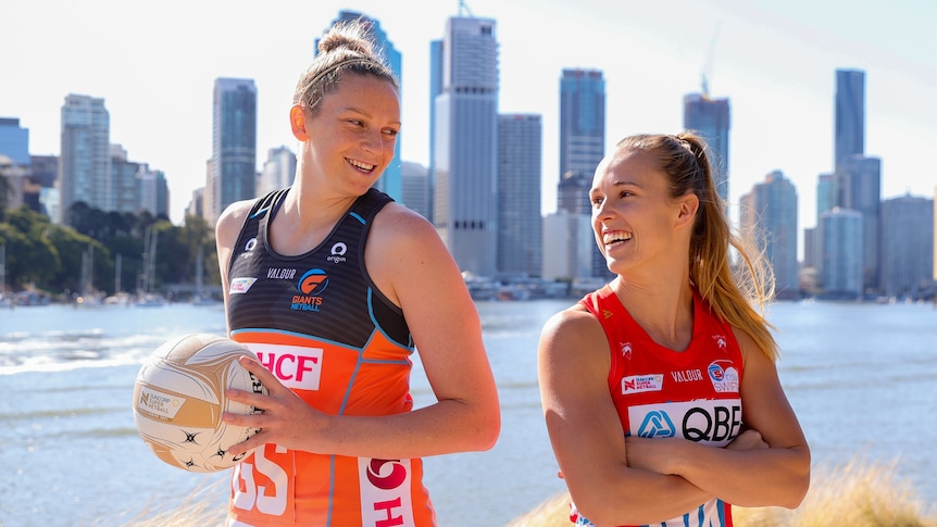 Jo Harten stands tall, holding the Grand Final ball, smiling back-to-back at Paige Hadley, in front of a Brisbane sky line
