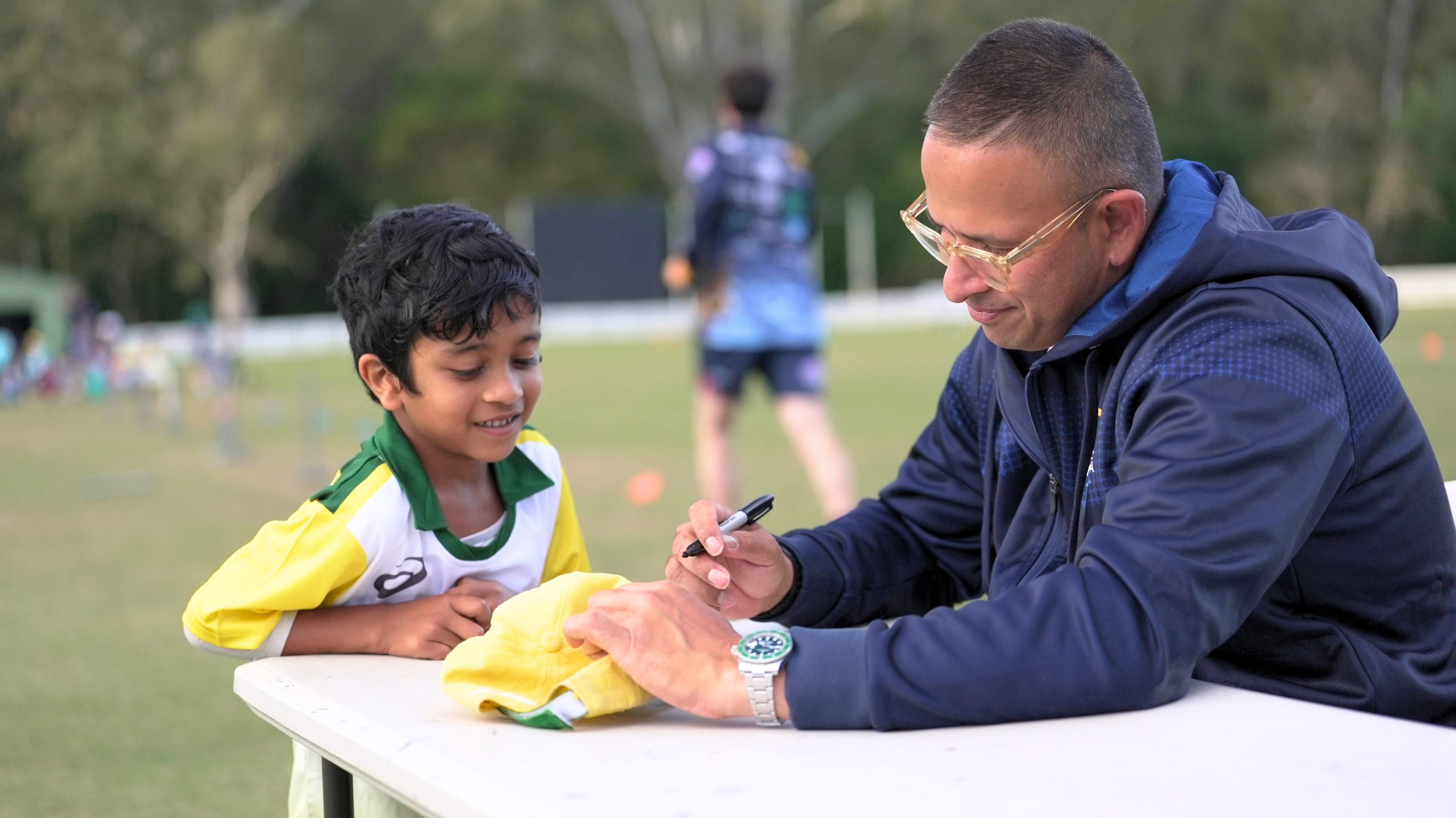 Usman Khawaja seated at a table outdoors wears glasses as he signs a cap for a young boy looking on smiling.