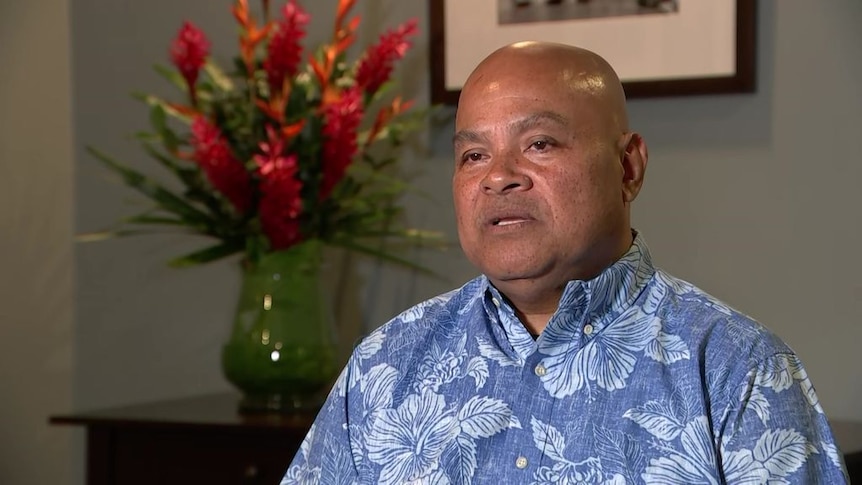 A bald man in blue and white shift sitting with flowers in background. 