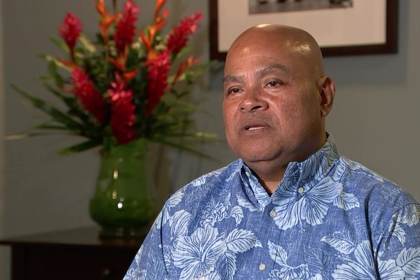 A bald man wearing a blue floral shirt being interviewed in a room decorated with flowers in background 