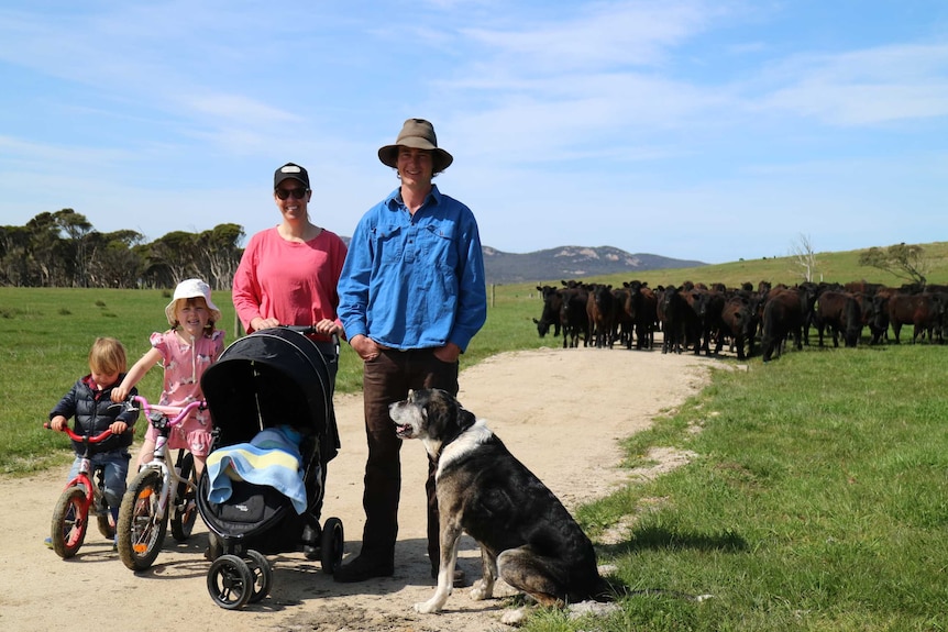 A family stands on a dirt road in the middle of their farm with a herd of cows in the background