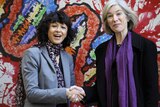 Two women smile as they shake hands while standing in front of a large colourful painting of a stylised DNA strand