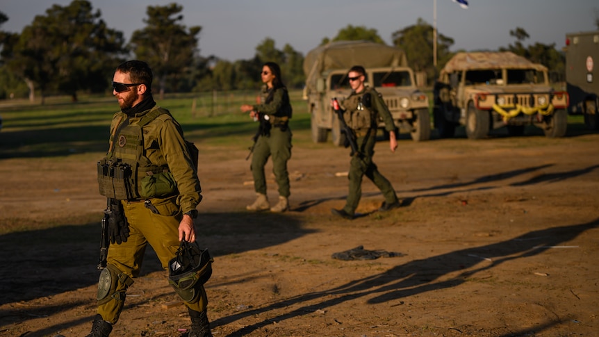 Three soldiers, two male, one female, walk near military vehicles and a flying Istael flag