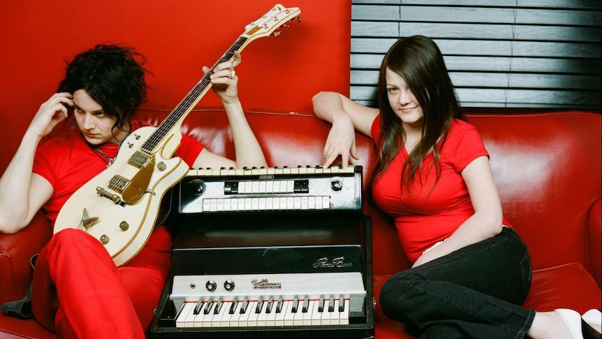 Jack and Meg White of The White Stripes sit on a red couch surrounded by instruments