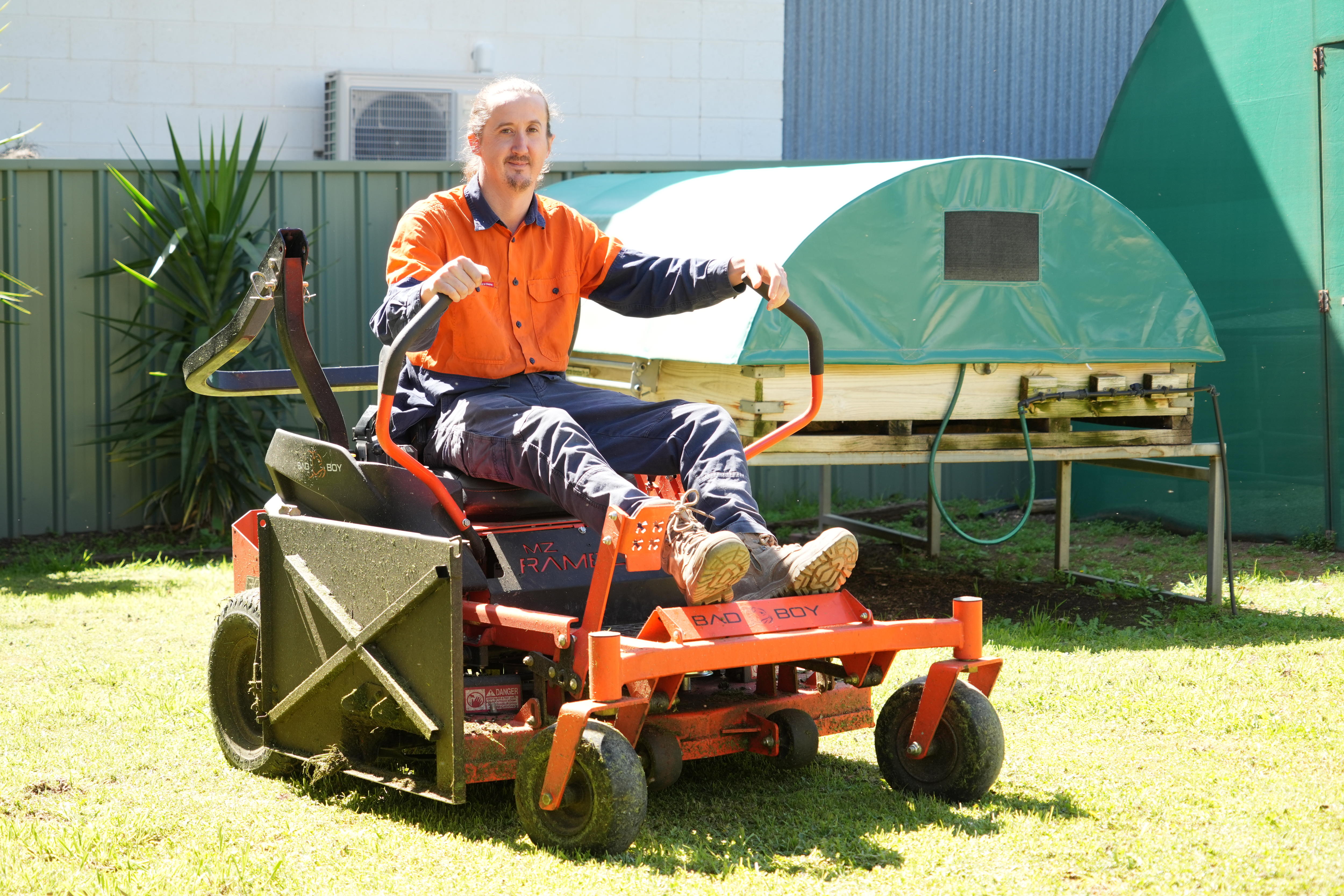 A man in an orange work shirt and pants sitting in a lawnmower on a patch of lawn