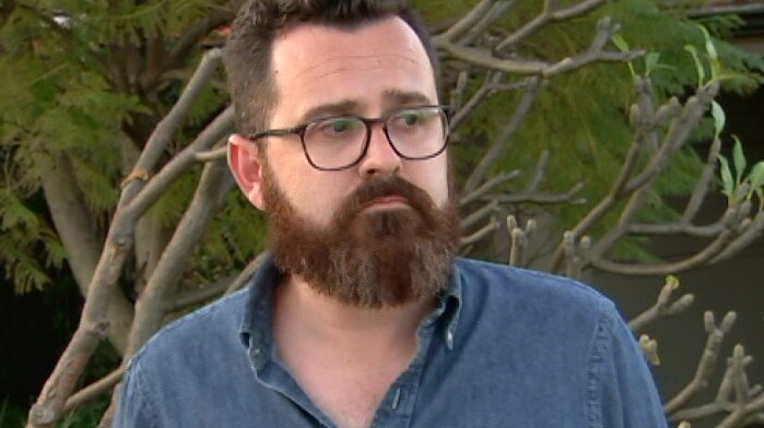 A man with a beard and glasses, wearing a blue shirt and standing in front of a tree.