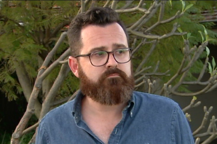A man with a beard and glasses, wearing a blue shirt and standing in front of a tree.