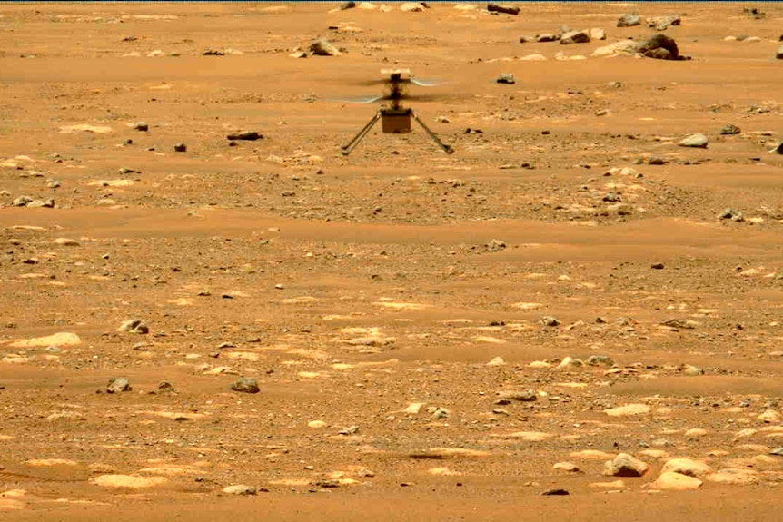 A small helicopter-like drown hovers above the surface of a dry, rocky plain. 