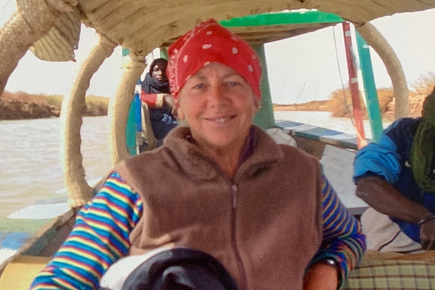 older woman wearing red bandanna with brown vest and colourful stripy shirt smiles at the camera while sitting on boat 