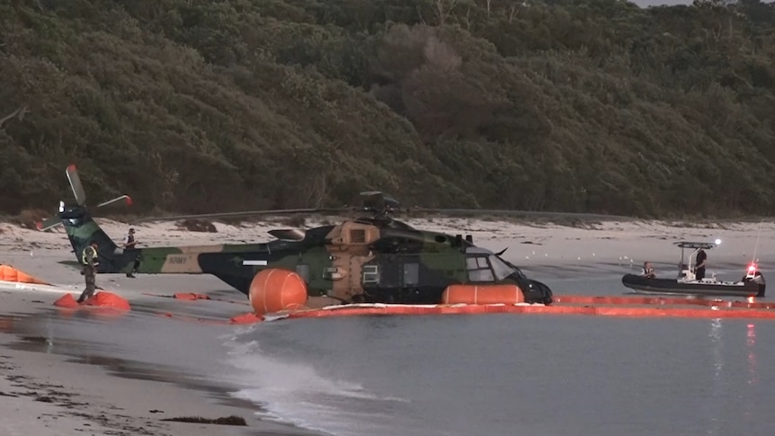 An image of an army helicopter in water near a beach as a small police boat approaches on the right side.